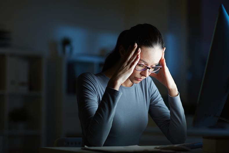  Stress, Depression ..Young businesswoman with headache touching her head while sitting in front of computer monitor by her workplace at night